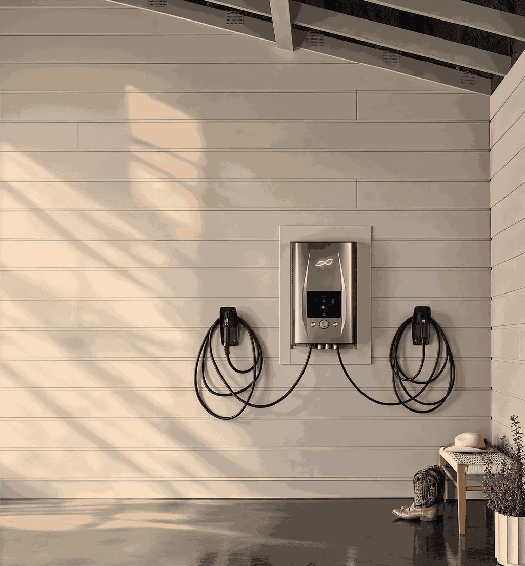 dcbel home energy station mounted on a white shiplap garage wall with two EV charging cables, sunlight filtering through exposed rafters onto a polished concrete floor with a bench, cowboy boots, and a potted plant in the corner.