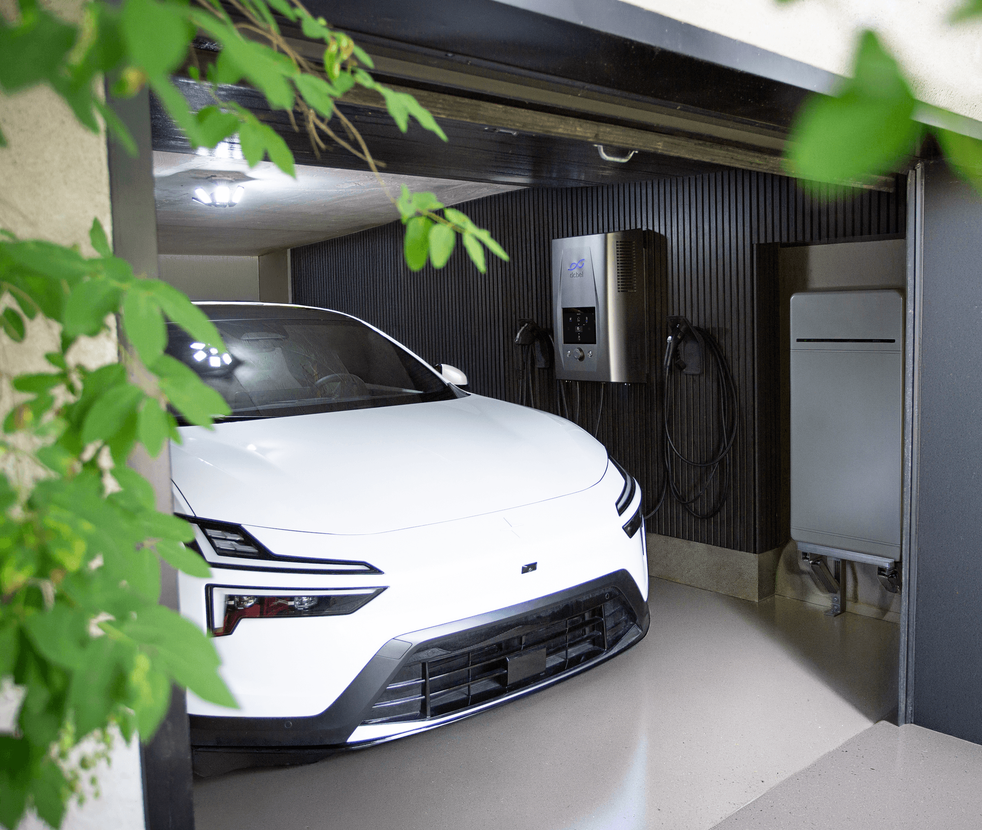 A white Polestar parked in a modern garage with dark slatted wood walls. On the back wall, a dcbel EV charger (silver unit with the dcbel logo) is mounted next to a home battery storage unit. Green foliage frames the left side of the image.